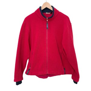 May include: A red fleece jacket with a full zipper closure and a navy blue collar and cuffs. The jacket has two side pockets with zippers and is hanging on a wooden hanger. The jacket is made of a soft, textured material.