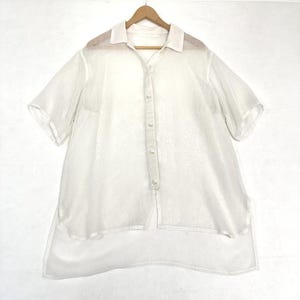 May include: A white, sheer button-down shirt with short sleeves, a collar, and a layered design. The shirt is hanging on a wooden hanger against a white background. The fabric appears lightweight and slightly translucent.