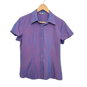 May include: A short-sleeved button-down shirt in a shimmering purple hue, hanging on a wooden hanger. The shirt features a classic collar and a series of small, round buttons down the front. The fabric has a subtle sheen, creating a dynamic visual effect.