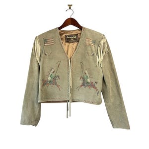 May include: A light olive green suede jacket featuring long cream-colored fringe on the shoulders. The front displays two painted illustrations of figures in headdresses riding horses, two American flags, and a label reading Patricia Wolf.