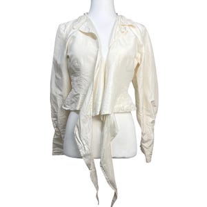 May include: Ivory-colored jacket with long sleeves and a draped front. The jacket features a collar and a tie closure, creating a flowing silhouette. The fabric has a subtle sheen, suggesting a silk or satin material. The garment is displayed on a mannequin.