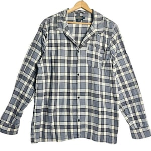 May include: A long-sleeved, button-down shirt in a gray, white, and black plaid pattern. The shirt has a single chest pocket and is hanging on a wooden hanger. The shirt is made of a soft, woven fabric.