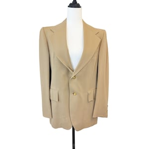 May include: A tan blazer with two gold buttons and two front pockets. The blazer is displayed on a white mannequin. The blazer has a classic design with notched lapels and a two-button closure. The sleeves have buttons at the cuffs.
