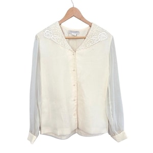 May include: Cream-colored blouse with a lace collar and sheer sleeves. The long-sleeved top has a button-down front with pearl-like buttons. The blouse is hanging on a wooden hanger.
