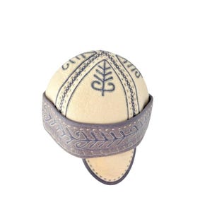May include: A dome-shaped hat made of cream-colored felt featuring dark blue embroidered tree-like motifs on the crown. The hat includes a wide, dark blue band around the base with intricate, repeating leaf-patterned embroidery and stitched edges.