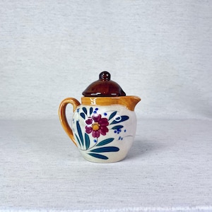 May include: A small, white ceramic teapot with a brown lid and floral design. The teapot features a pink flower with green leaves and blue accents.