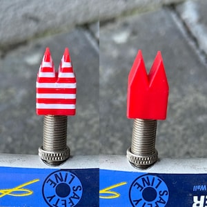 May include: Two bicycle valve caps. One is red and white striped, the other is solid red. Both have a distinctive, angular design. They are mounted on a silver threaded stem, likely for a bicycle tyre.