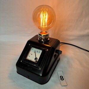 May include: A retro desk lamp with a large, round Edison bulb. The lamp has a black, angled base with a voltmeter and a knob. The bulb emits a warm, orange glow, and a black power cord extends from the base.