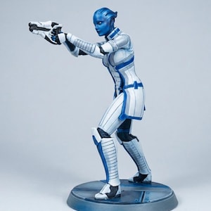 Puede incluir: Una figura de piel azul de un personaje femenino con un traje blindado blanco y azul, sosteniendo un arma blanca y negra. La figura está posada en una postura dinámica sobre una base circular, con armadura y rasgos faciales detallados.