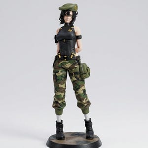 Op de afbeelding: Een beeldje van een vrouw in militaire kleding. Ze draagt een groene baret, een zwarte top, een camouflagebroek en zwarte laarzen. Een kleine groene tas hangt aan haar riem. Het beeldje staat op een donkere, ronde basis.