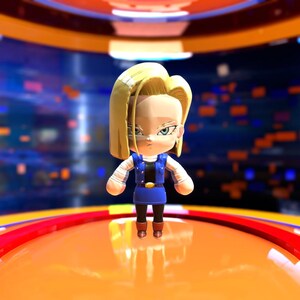 May include: A stylized figurine of Android 18 from Dragon Ball Z. The figure has blonde hair, blue eyes, and is wearing a blue outfit with a black vest. The figure is standing on an orange platform with a blurred background.