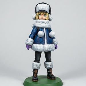 May include: A figurine of a person in winter attire. The figure wears a dark blue coat with white trim, a matching hat, and black pants. The figure is standing on a green base. The figure has blonde hair and green eyes.