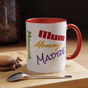 May include: White ceramic mug with a red interior and handle. The mug features the words "Momma", "Mum", "Mommy", and "Madre" in various colors. A spoon and a macaron are on a wooden surface.