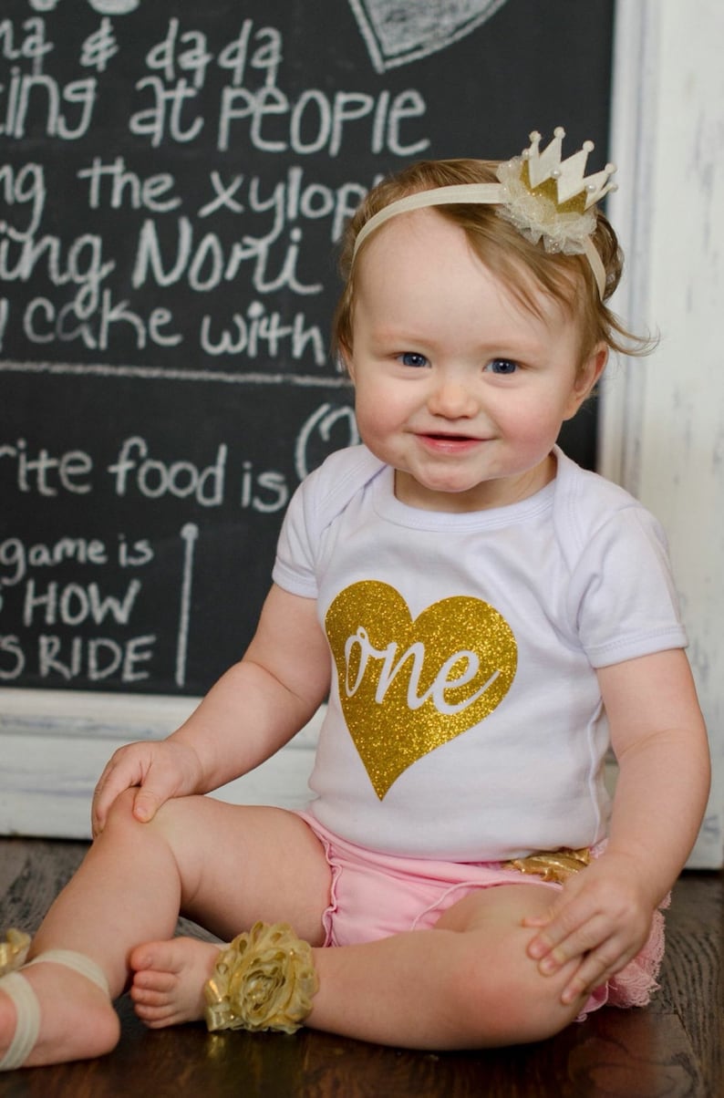 May include: A baby wearing a white onesie with a gold glitter heart that says "one". The baby is also wearing a gold crown headband and pink ruffled shorts. The background is a chalkboard with white writing.