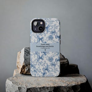 May include: A phone case with a floral and bow pattern in blue and white. The case has a light blue band with the text "Love over Knowledge and Faith" and a Bible verse reference. The case is on a stone surface.