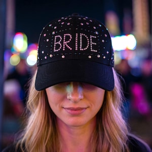 Peut inclure: Casquette de baseball noire avec le mot "BRIDE" en strass roses. La casquette est ornée de strass supplémentaires. L'image est prise de nuit avec des lumières floues en arrière-plan.