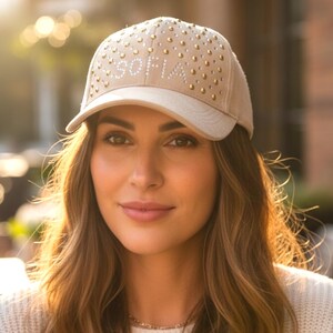 Peut inclure: Casquette de baseball beige avec le nom "SOFIA" en lettres blanches et des clous dorés. La personne sur l'image porte un pull blanc. Un accessoire de mode.