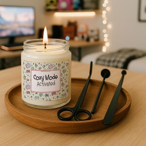 以下が含まれることがあります： 木製のトレイに「Cozy Mode Activated」の文字が入ったガラス瓶に入った点灯したキャンドル。ハサミや消火器など、黒いキャンドルアクセサリーがキャンドルの横に置かれています。背景はぼやけています。