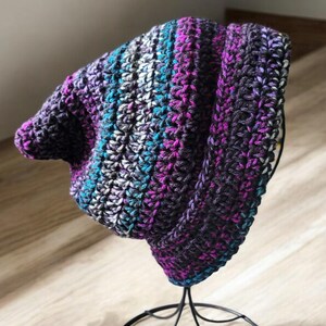 Può includere: Un cappello a beanie lavorato a uncinetto con strisce di nero, viola, verde acqua e bianco.