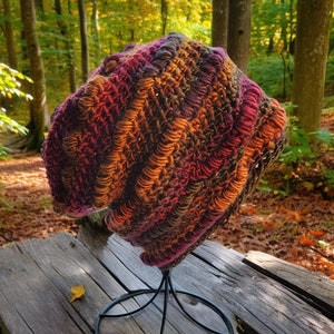 May include: A crocheted beanie hat in shades of brown, orange, and purple. The hat is displayed on a black metal stand.