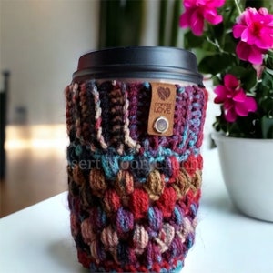 May include: A crocheted coffee cup cosy with a brown leather tag that says "Coffee Love". The cosy is made with a variety of colours, including red, brown, blue, and purple.
