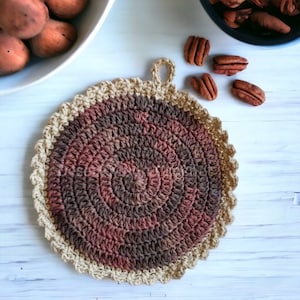 May include: A round crocheted pot holder with a brown, pink, and beige color scheme. The pot holder has a loop handle and a scalloped edge.