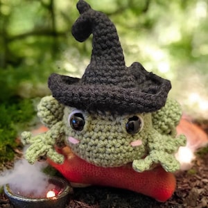 May include: A crocheted green frog wearing a black witch hat. The frog has large black eyes, pink cheeks, and is sitting on a red mushroom. A small cauldron with smoke is in the foreground, suggesting a Halloween theme.