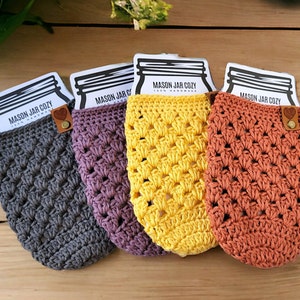 May include: Four handmade crochet mason jar cozies in gray, purple, yellow, and orange. Each cozy has a decorative pattern and a leather tag with a button. The top of each cozy has a card that reads "MASON JAR COZY 100% HANDMADE".