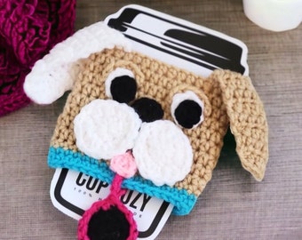 Op de afbeelding: Een gehaakte, hondvormige koffiebekerwarmer met bruine, witte en roze accenten. De bekerwarmer ligt op een houten oppervlak met een koffiebeker en een melkcontainer op de achtergrond.