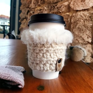 Pode incluir: Capa de crochê branca para caneca de café com acabamento em pele sintética e uma tira de couro marrom com um logotipo de coração. A capa está em uma caneca de café branca com uma tampa preta.