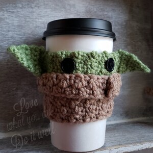 May include: A green and brown crocheted coffee cup cozy shaped like a character from Star Wars. The cozy has two black buttons for eyes.