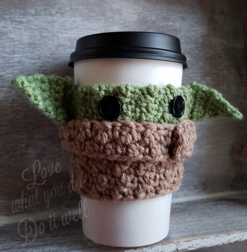 May include: A green and brown crocheted coffee cup cozy shaped like a character from Star Wars. The cozy has two black buttons for eyes.