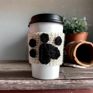 May include: A white paper coffee cup with a black lid is wrapped in a cream-colored, crocheted cozy. The cozy features a black paw print design. The cup is on a wooden surface.