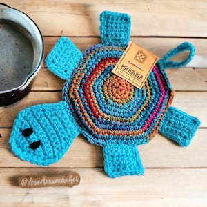 May include: A handmade, crocheted turtle-shaped pot holder. The turtle features a blue head, flippers, and tail, and a shell in shades of blue, orange, red, and pink. A tag reads "POT HOLDER".