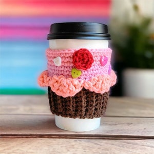Pode incluir: Um protetor de copo de café de crochê rosa e marrom em forma de cupcake com botões rosa, branco e verde.