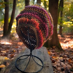 May include: A crocheted beanie hat in shades of brown, pink, and orange, displayed on a black metal stand. The hat is made with a textured, chunky yarn and has a slouchy fit.