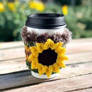 Pode incluir: Um aquecedor de xícara de café de crochê com uma borda de pele sintética marrom e um design de girassol amarelo.