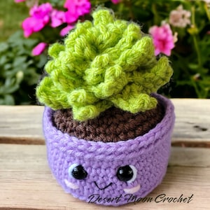 Pode incluir: Uma planta suculenta de crochê em um vaso roxo com um rosto bonito. O vaso tem terra de crochê marrom e a suculenta é verde.