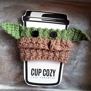 May include: A green and brown crocheted cup cozy shaped like a character from Star Wars. The cozy has two black buttons for eyes and is designed to fit a standard-sized cup. The text "CUP COZY 100% HANDMADE" is printed on a white label.
