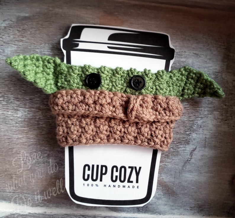 May include: A green and brown crocheted cup cozy shaped like a character from Star Wars. The cozy has two black buttons for eyes and is designed to fit a standard-sized cup. The text "CUP COZY 100% HANDMADE" is printed on a white label.