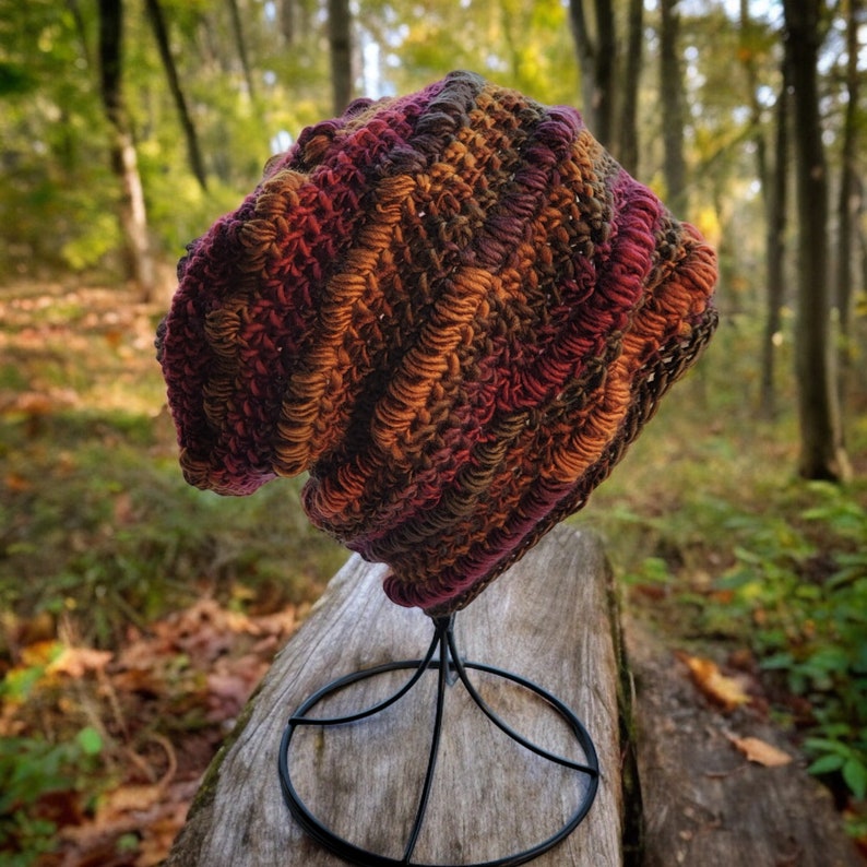 May include: A crocheted beanie hat in shades of brown, red, and orange. The hat is displayed on a black metal stand.