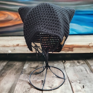 May include: A black crocheted cat ear beanie hat with a brown leather tag. The hat is displayed on a black metal stand.