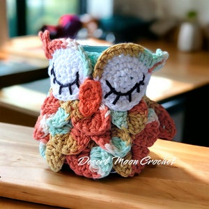 Peut inclure: Un dessous de verre en crochet fait à la main en forme de hibou aux couleurs pastel. Le hibou a des yeux blancs avec des cils noirs, des accents orange et jaune, et un design texturé et superposé. Le texte "Desert Moon Crochet" est visible.