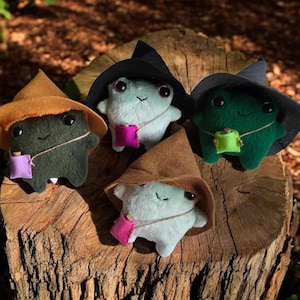 Puede incluir: Cinco figuras de rana de peluche con caras sonrientes, que llevan sombreros de bruja y pequeñas bolsas. Las ranas son de varios colores: verde, negro y blanco. Los sombreros son marrones y negros. Las bolsas son rosas, moradas y verdes. Están dispuestas sobre un tocón de árbol.