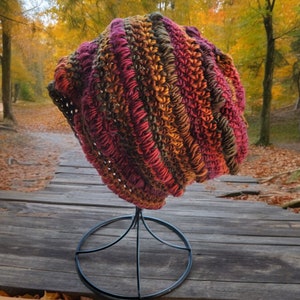 May include: A crocheted hat in shades of red, orange, brown, and green. The hat is displayed on a black metal stand.