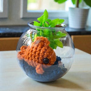 May include: A clear glass fishbowl contains a crocheted orange fish, blue pebbles, and green plastic plants. The fish has a large black eye. The bowl sits on a light-coloured surface, with a blurred background.