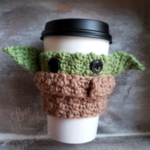 May include: A crocheted coffee cup cozy in the shape of a green alien character with brown ears and black button eyes. The cozy is on a white coffee cup with a black lid.