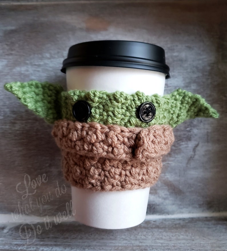 May include: A crocheted coffee cup cozy in the shape of a green alien character with brown ears and black button eyes. The cozy is on a white coffee cup with a black lid.