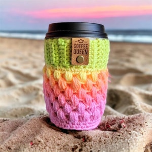 Pode incluir: Um aconchegante de xícara de café de crochê colorido com uma etiqueta de couro marrom que diz "Coffee Queen". O aconchegante é em tons de verde, amarelo, laranja e rosa. O aconchegante está sentado em uma praia de areia.