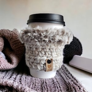Pode incluir: Um aquecedor de xícara de café de crochê branco com uma borda de pele sintética. O aquecedor tem uma etiqueta de couro marrom com a palavra "LOVE" nela.